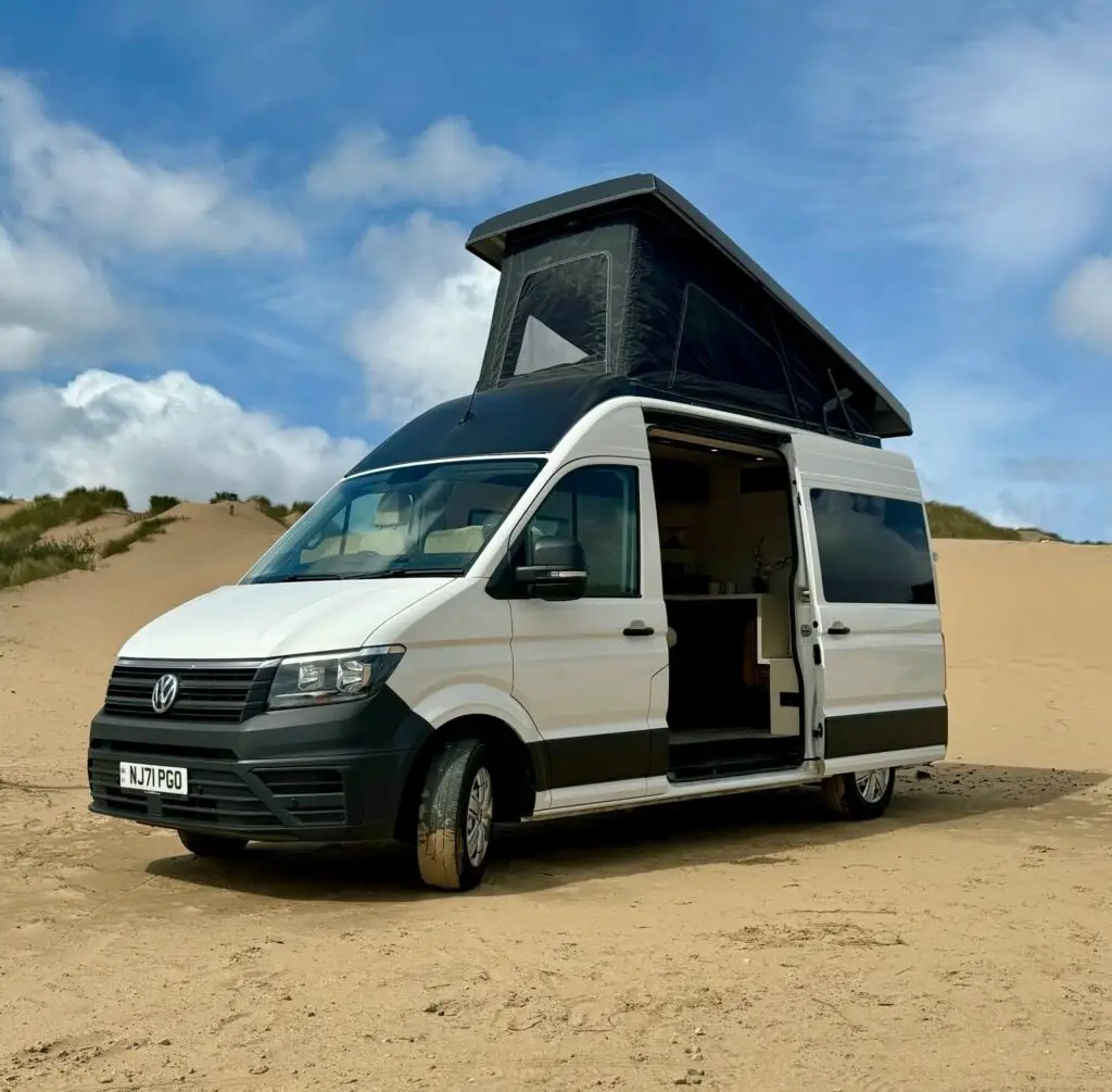 Campervan conversion with pop-top roof extended and side door open to reveal living interior. The campervan is parked on a sandy beach